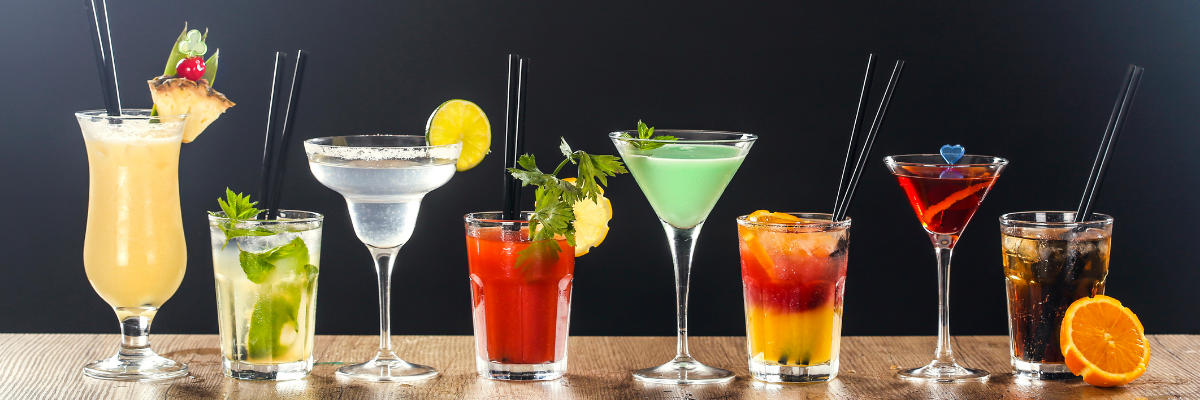 A variety of cocktails lined up in different glasses ranging from margaritas, daiquiris, martinis and classic cocktails on a wooden table with a black background