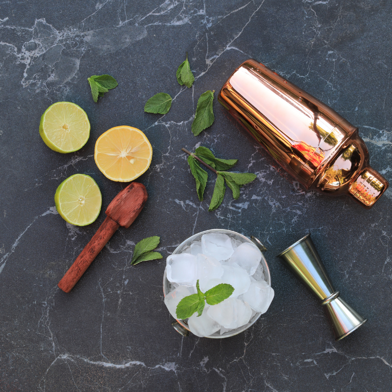 Cocktail shaker, jigger, muddler, ice, lemons, limes and herbs flat laid on a marble counter