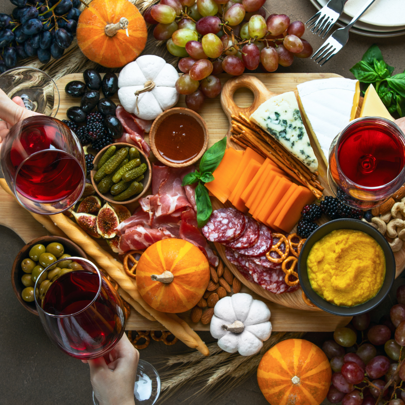 Charcuterie board with cheese, mustard, honey, cured meats, grapes, pickles, figs and other items on a table with two people's hands holding glasses of red wine  